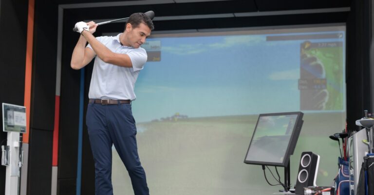 A man practicing his golf game in a professional indoor golf simulator system. He is checking his form on a monitor.