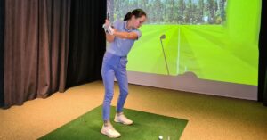 A young and experienced golfing enthusiast lining up her driving swing in front of an indoor golfing simulator machine.