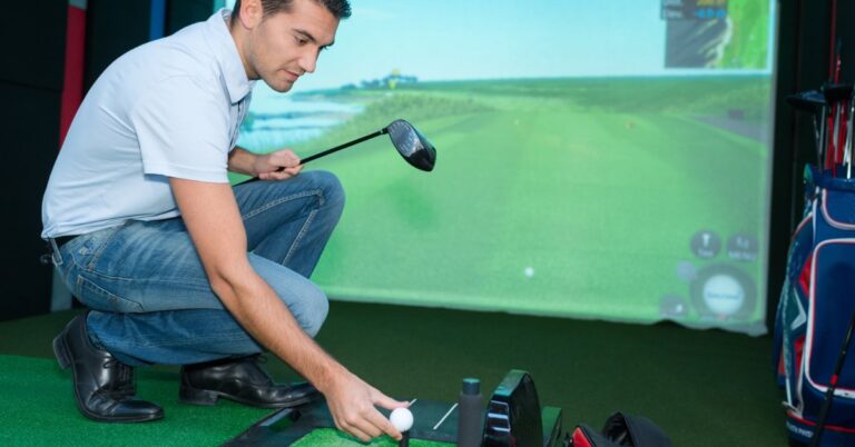 A man placing a golf ball on the tee of an indoor golfing simulator machine while holding his driving club in the other hand.
