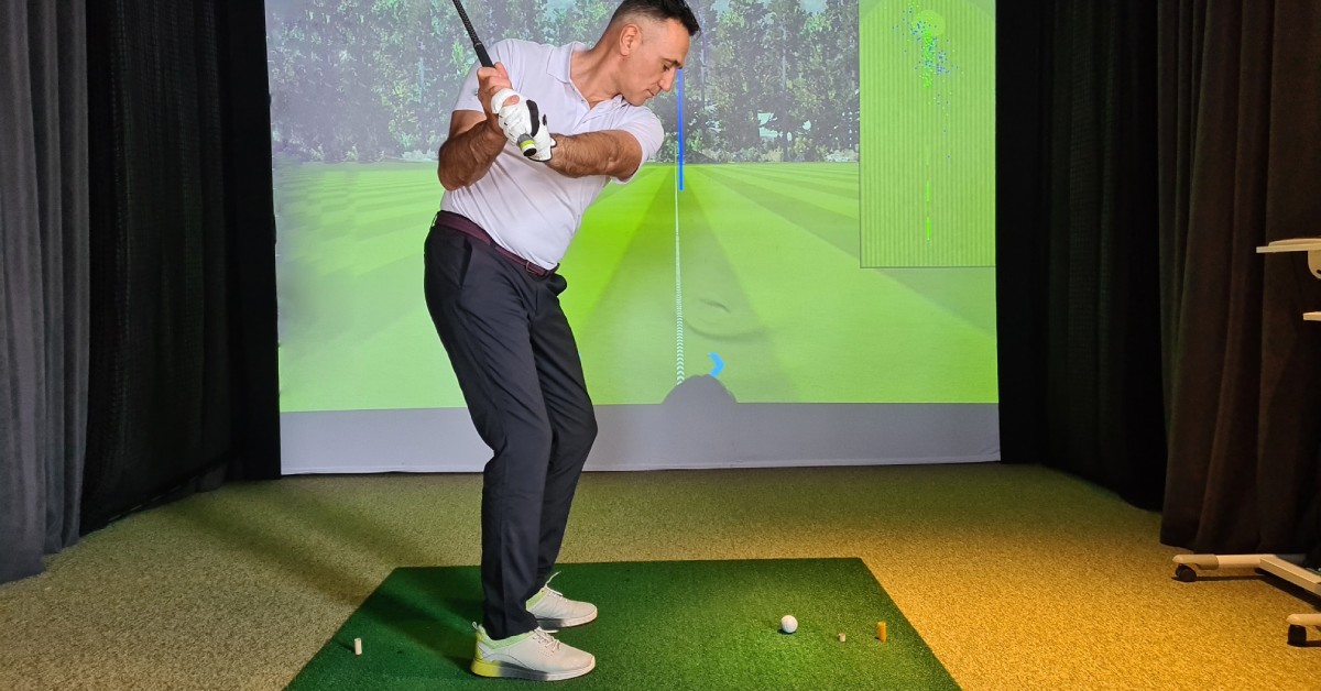 A male golfing enthusiast in the middle of his driving swing and aiming towards a virtual golf simulator.